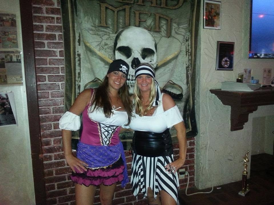 Woody's Pirate Party — two women in pirate wench costumes posing in front of Dead Men skull and crossbones flag inside Woody's bar