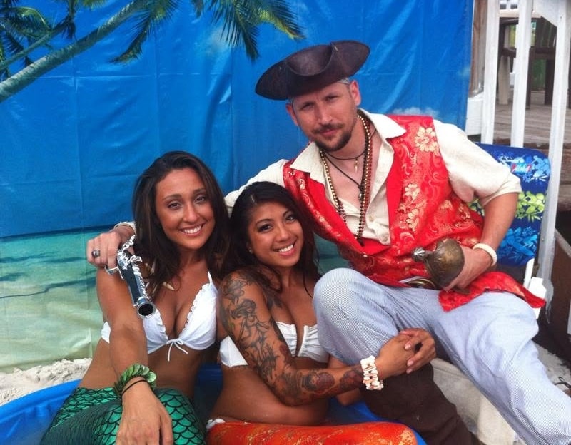 Woody's Pirate Party — man in red brocade vest with two women in white bikinis at tropical beach backdrop