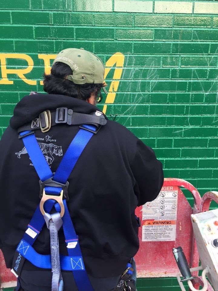 Close-up of crew member hand-lettering the yellow "We Are the Mighty Salt City" text on the green field