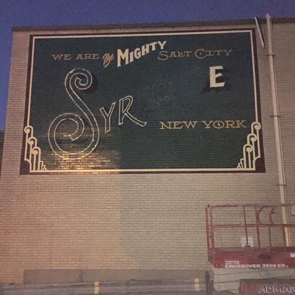 Night shot — "Syracuse" lettering partially painted in under artificial light, mural in final stages