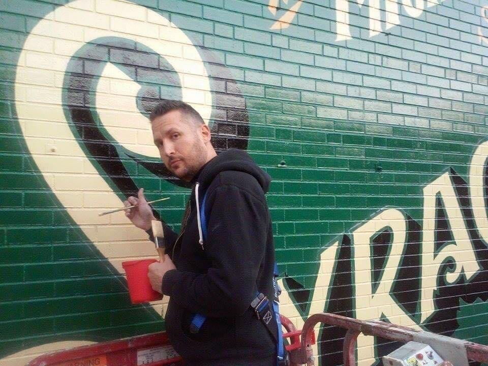 Marcus Osmun on the scissor lift, brush and paint cup in hand, looking back at camera